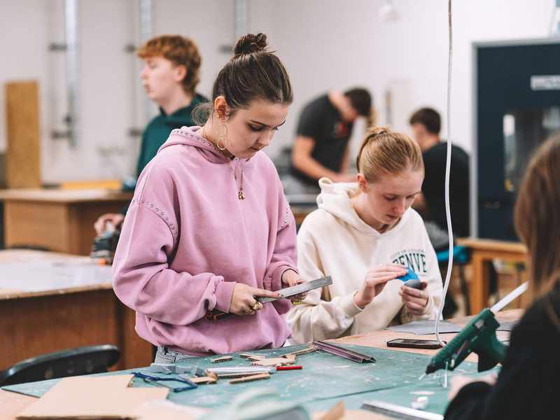 Students working at a table cutting materials and assembling small card models in a workshop.