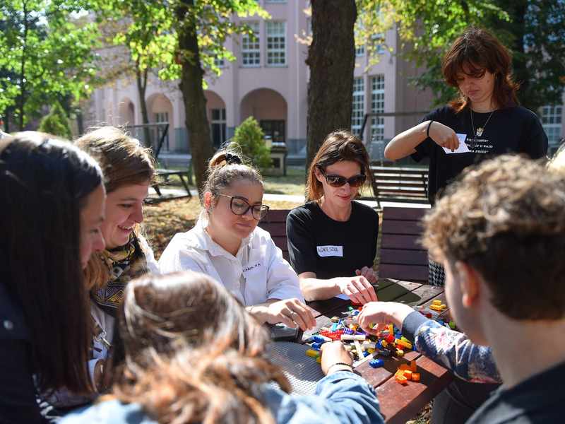 A group of people wearing name badges sit around a table outdoors. On the table in front of them are LEGO bricks.