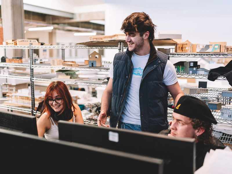 Students gathered around computer workstations in a bright studio space with architectural models displayed on shelves behind.