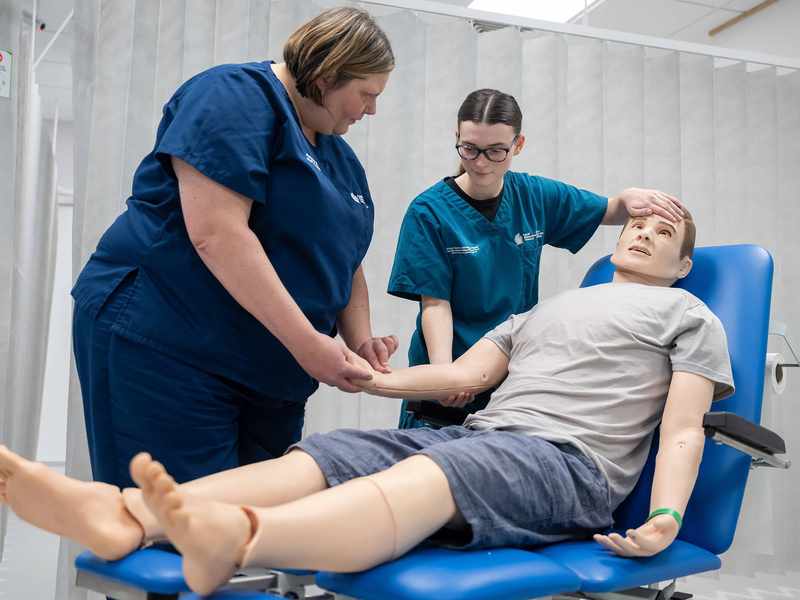 A lecturer demonstrates how to measure a patient's pulse using a manikin.