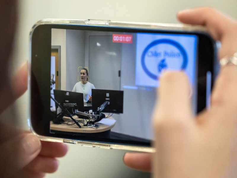 A close-up of a smartphone screen recording a video. On the screen is a person sat at a desk in front of two computer monitors.