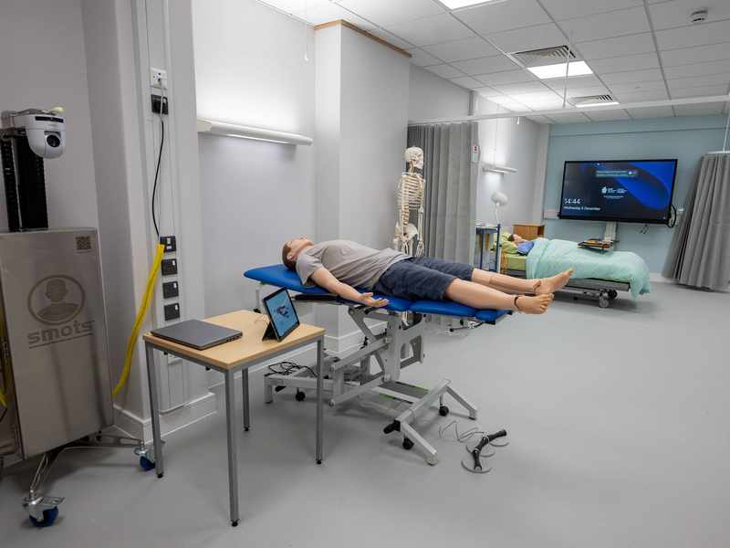 A manikin lies on a height-adjustable trolley in a simulated clinical environment.