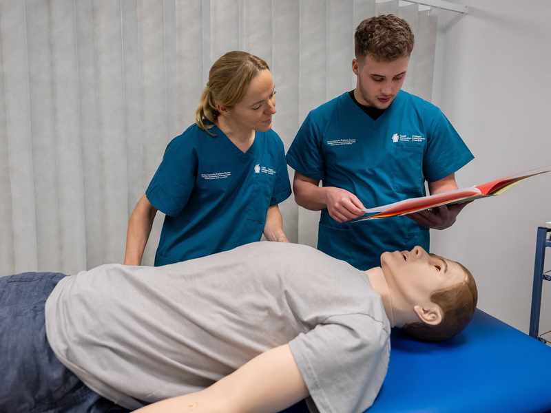 A manikin lies on a trolley in a simulated clinical environment, while a student consults patient records.