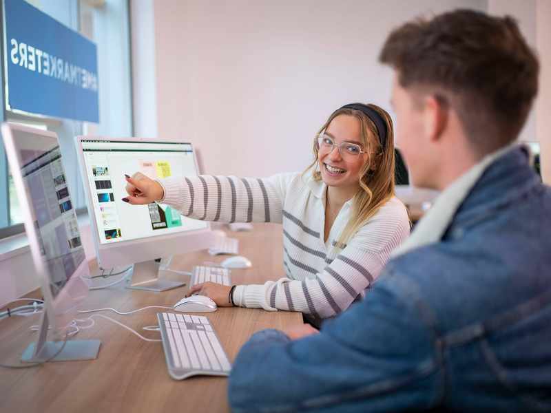 A desk with two people working on computers. One person is pointing to a computer screen while turning to face the other person.