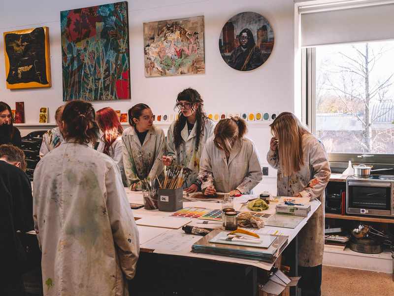 A group of students gather around a large table on which there are brushes and other painting materials. The students are wearing white coats spattered with paint.