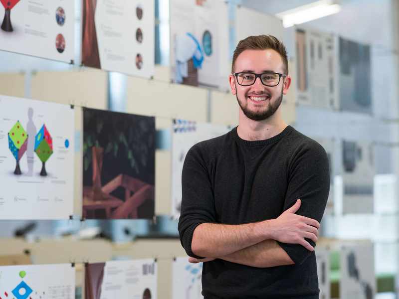 A young man in a dark sweater stands with his arms folded. Behind him is a display of drawings and designs.
