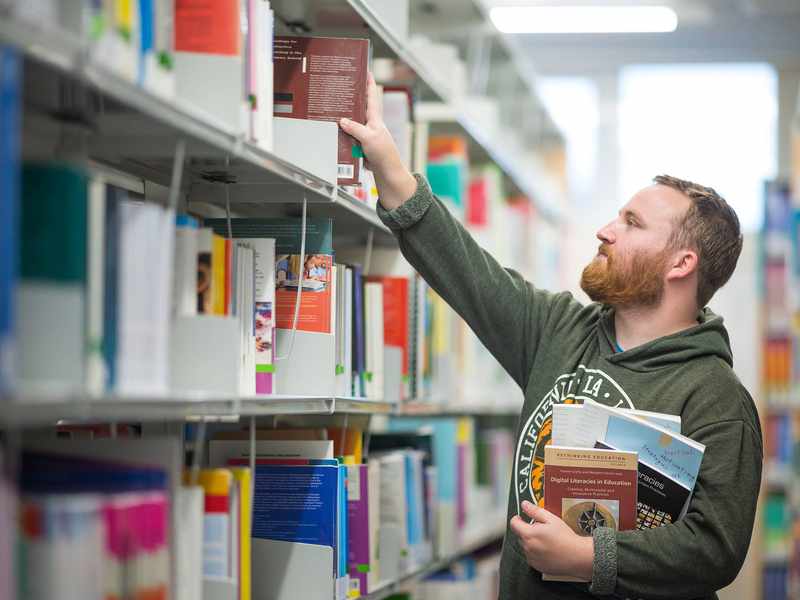 A young man with a beard reaches for a book on a library shelf, while holding several other books underneath his arm.