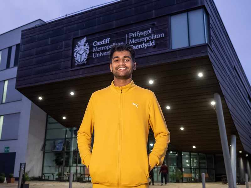 A young man in a yellow sweatshirt stands in front of a large modern building.
