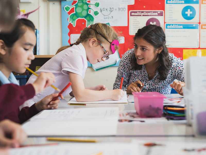 A school teacher sits at a table next to two young children. The children are writing in notebooks.