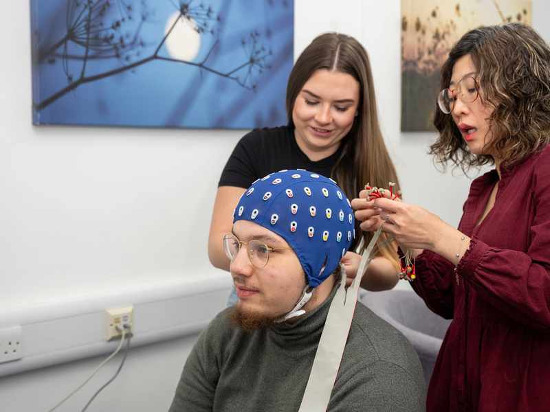 Two people assist a seated person with an EEG cap. One adjusts the cables on the cap, while the other looks on.