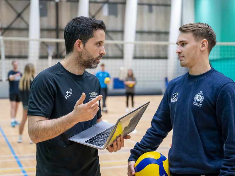 Two people converse in a gym. One holds a laptop computer, while the other holds a volleyball.