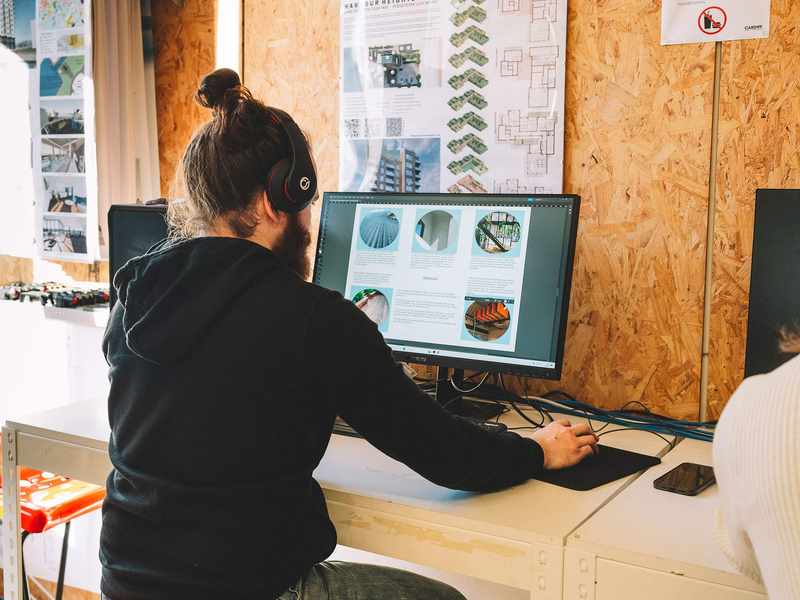 Student using a desktop computer at a workstation with architectural posters on the wall.