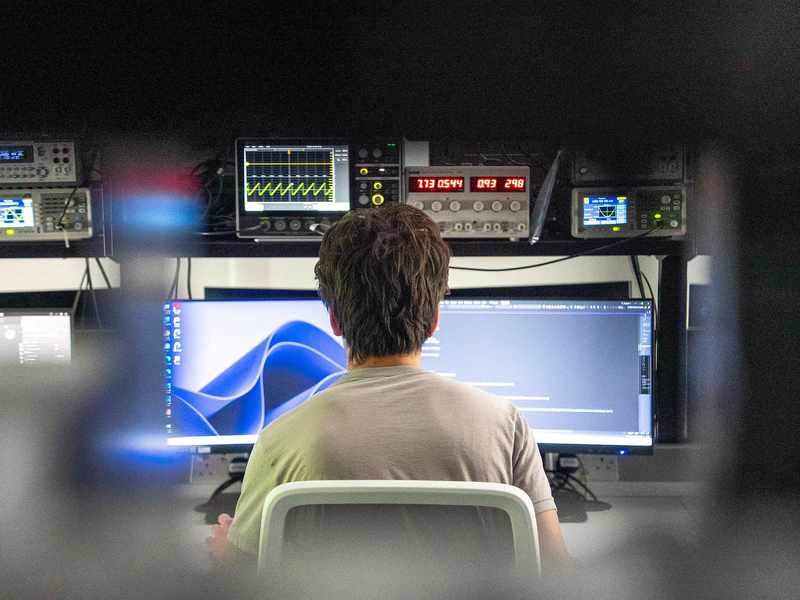 Student facing two computer monitors and a rack of electronic equipment