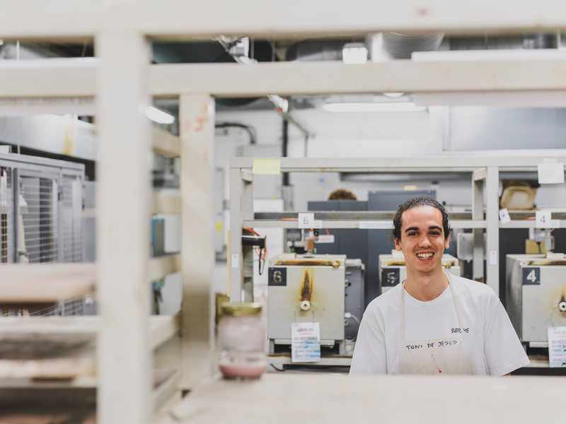 A young man stands in a ceramics workshop. Behind him is a row of small kilns.
