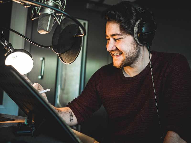 A man wearing headphones smiles while sitting in front of a microphone in a recording studio. He holds a pen and appears to be working or recording audio. The setting is dimly lit, with a lamp illuminating his workspace.