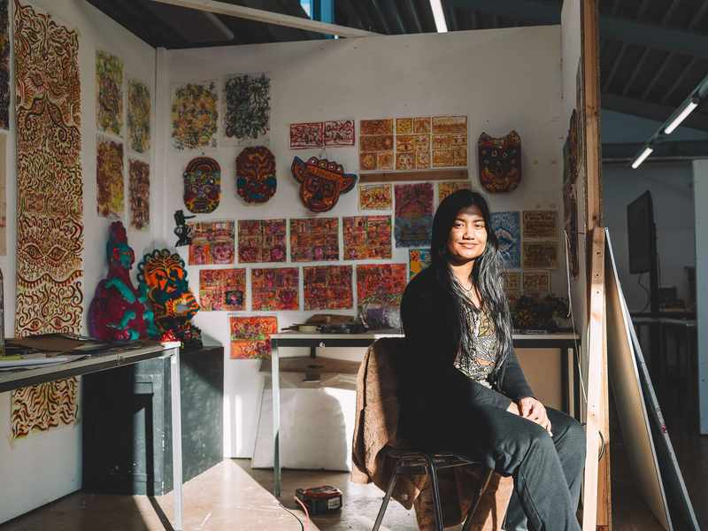 A student sits in front of a wall filled with various pieces of colourful artworks.