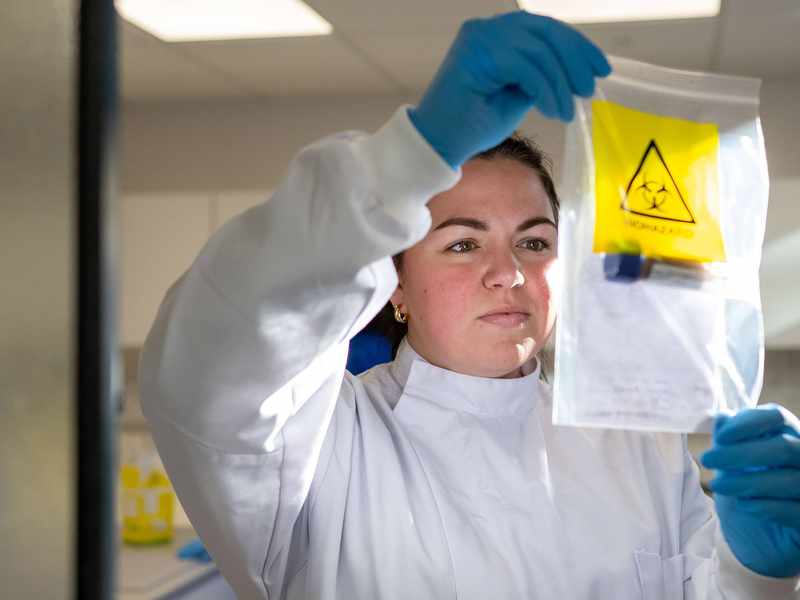 Student in laboratory coat holds clear bag containing test tube