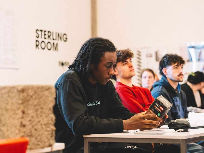 Student holding a boxed product while seated at a classroom table with others listening nearby.