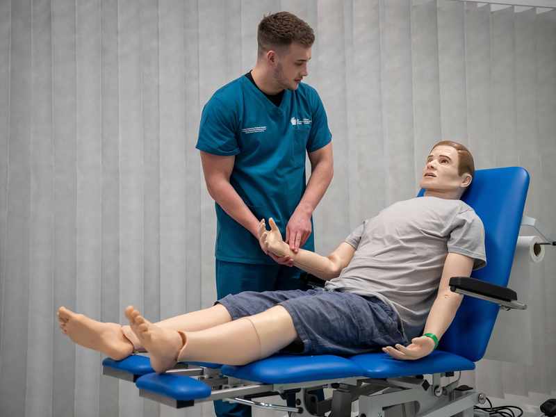 A student takes the pulse of a manikin lying on a height-adjustable trolley in a simulated clinical environment.