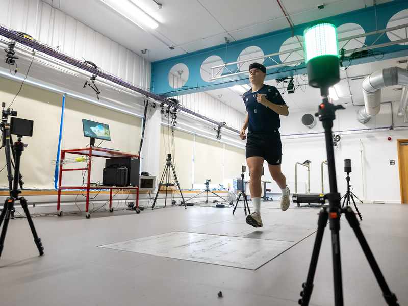 A person runs in a technology-equipped lab, surrounded by cameras and a computer screen. A bright green light is visible in the foreground. The setting suggests a motion analysis or biomechanics study.