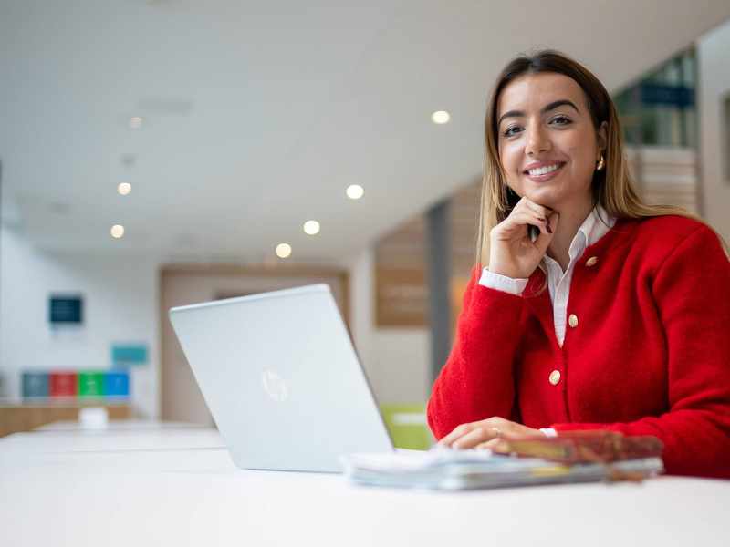 A person is smiling while sitting at a table with a laptop in front of them. They are in a bright, modern setting.