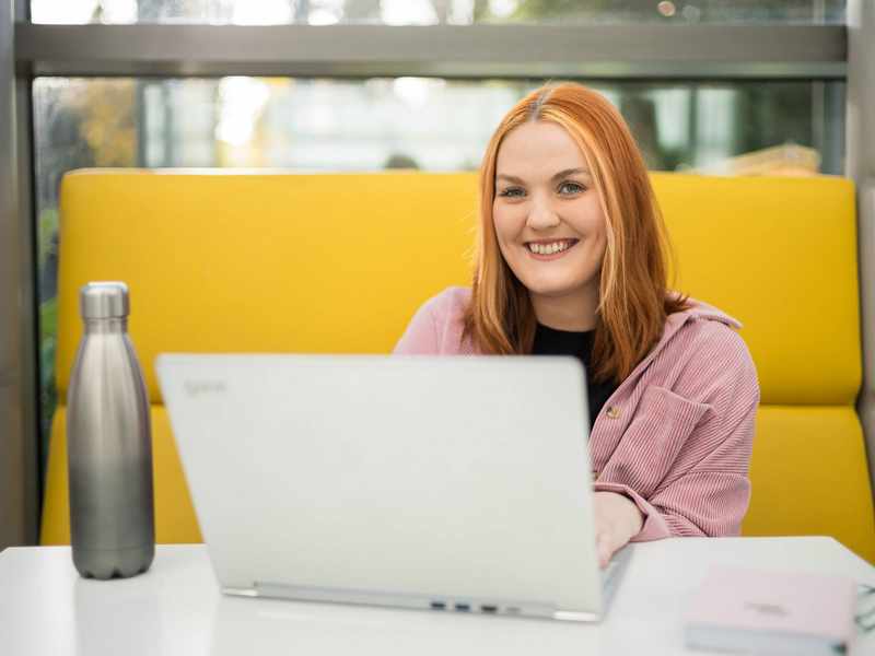 A young woman is smiling while sitting at a table with a laptop in front of them. They are in a bright, modern setting, with a yellow cushioned seating area behind her.