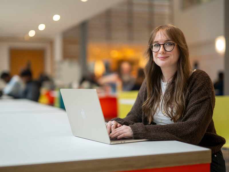 A person is smiling while sitting at a table with a laptop in front of them. They are in a bright, modern setting.