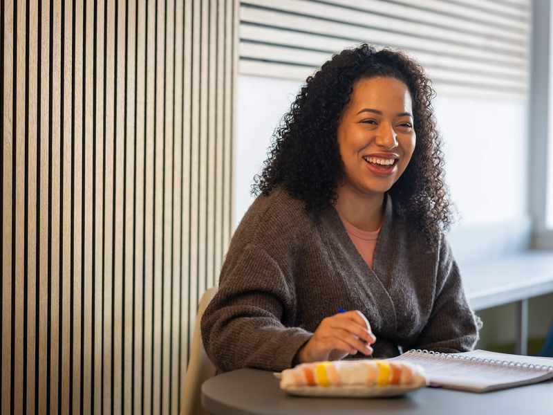 A young woman sits and smiles at a table while working with a pen and pad of paper.