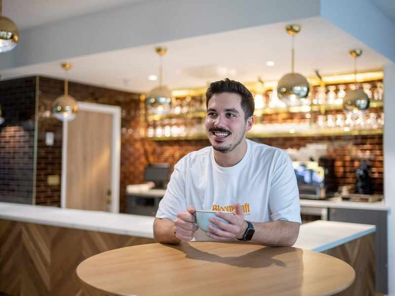 A young man sits at a table holding a coffee cup. Behind him is a bar area with racks of glasses.