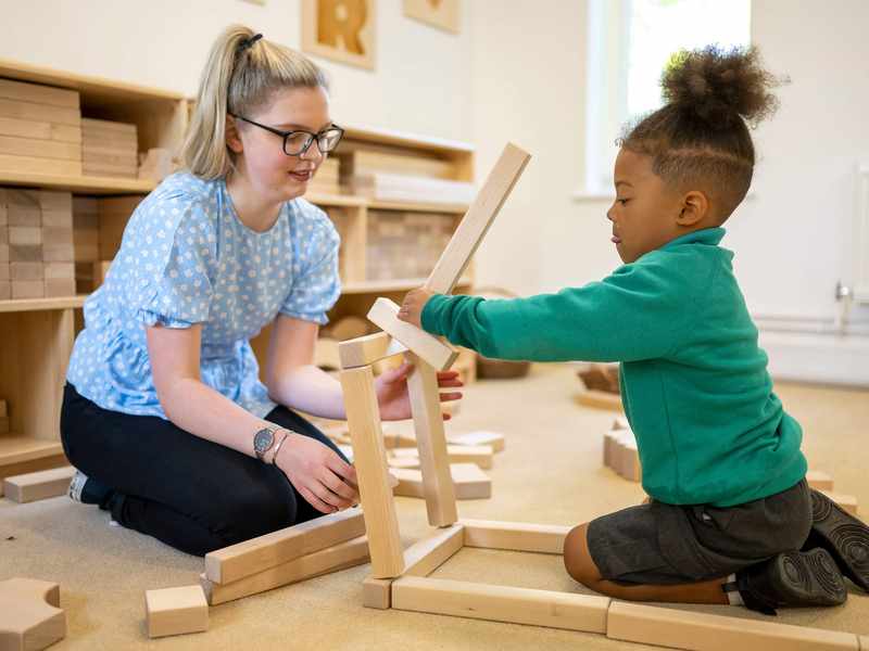 A child plays with wooden blocks as a teacher looks on.