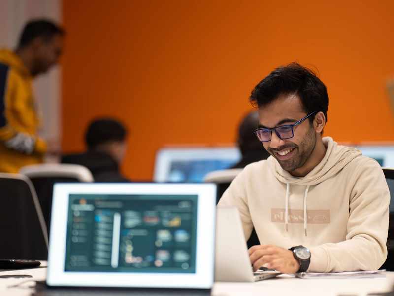 A student wearing a white hoodie and glasses is focused on his laptop while working at a desk.