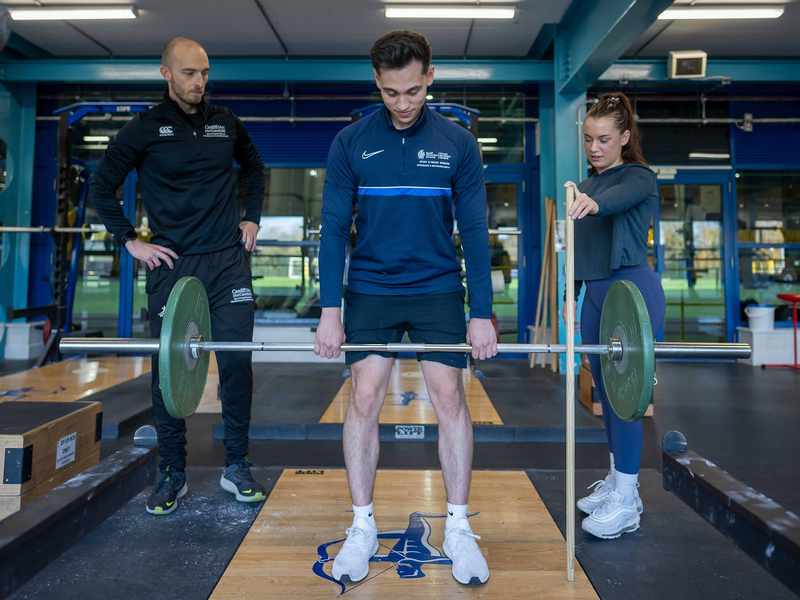 A person lifts a weight in a gym while two other people watch.