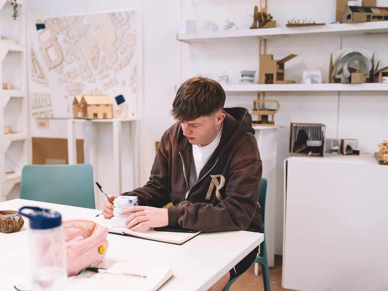 Student sitting at a desk writing in a notebook with architectural models displayed on shelves behind.