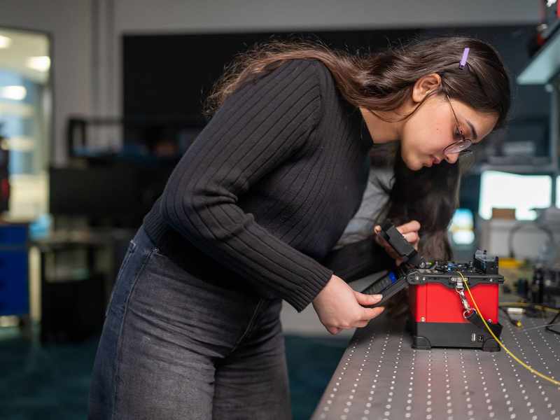 A student pressing the touchscreen of a small electronic device. In the background are other devices and computer monitors.