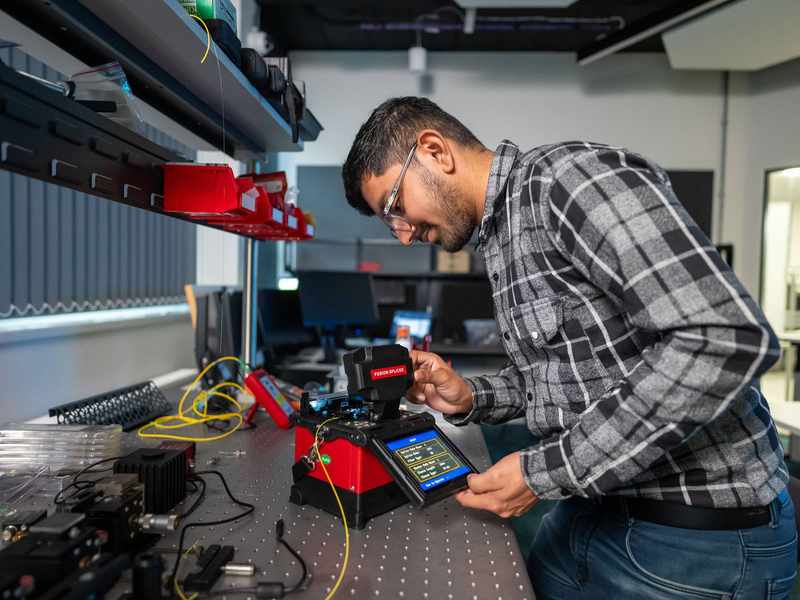 A student pressing the touchscreen of a small electronic device. In the background are other devices and computer monitors.