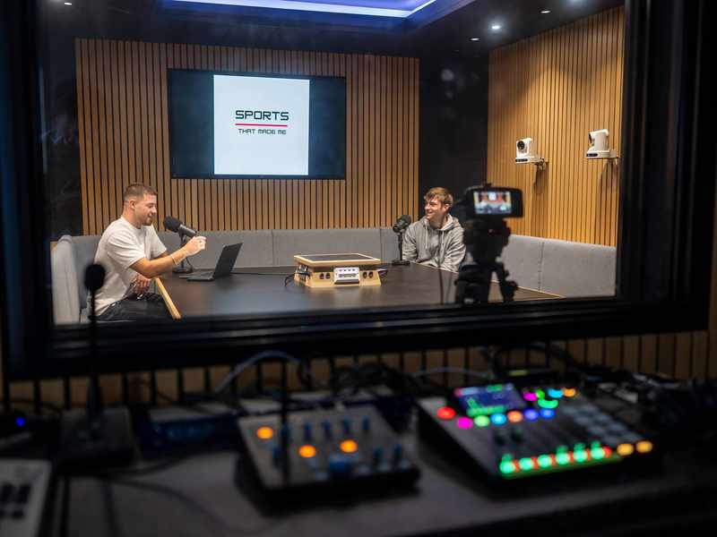 Two people sit at a table in a sound booth talking to one another. In front of them are microphones and other audio equipment.