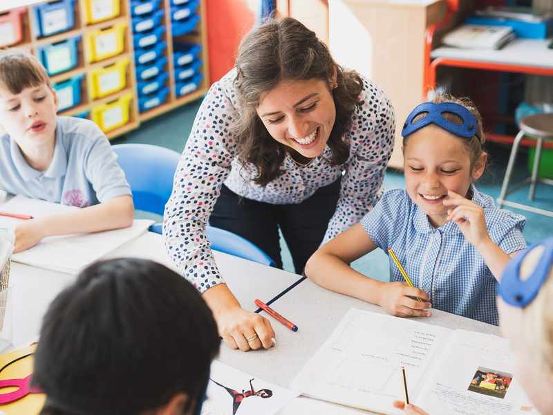 A teacher between two pupils sitting at a desk. The teacher is looking at an open book in front of one of the pupils.