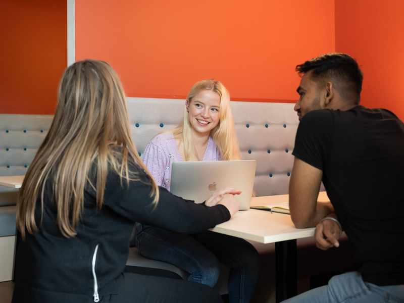Three students sit together on bench seating at a table. The wall behind them is a vibrant orange red.