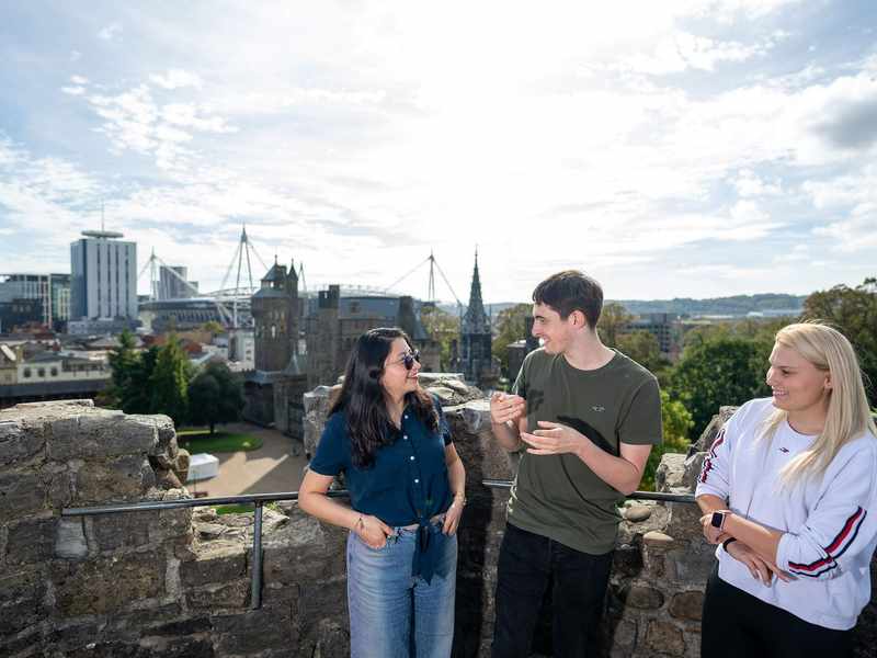 Three people converse while standing on the wall of a castle, with a panoramic view of a city in the background.