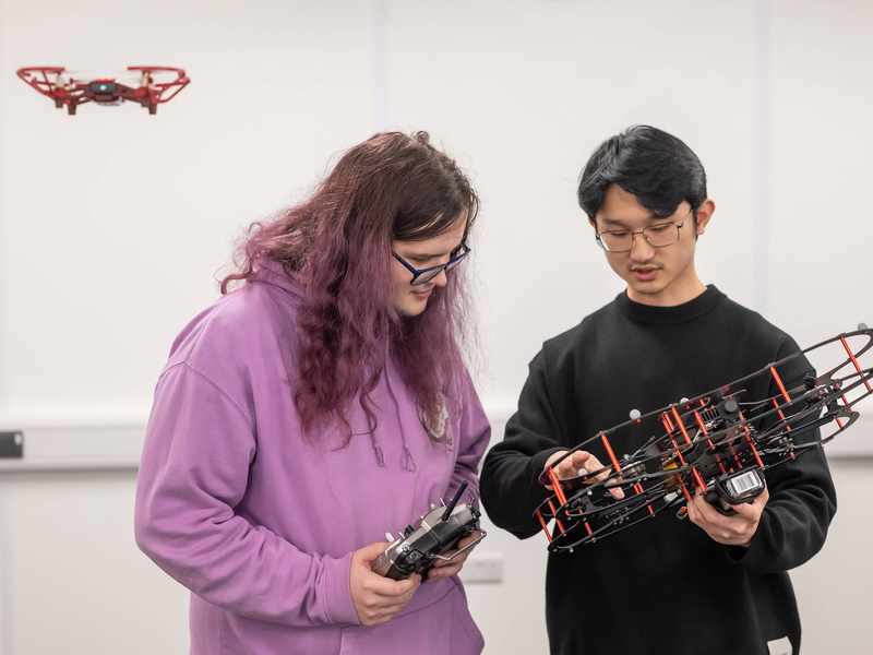 A person holding a drone quadcopter points to one section of it. A second person holding a remote control looks on. In the background a smaller quadcopter hovers in the air.