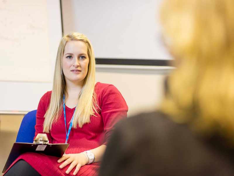A woman wearing a lanyard with the NHS logo holding a clipboard.
