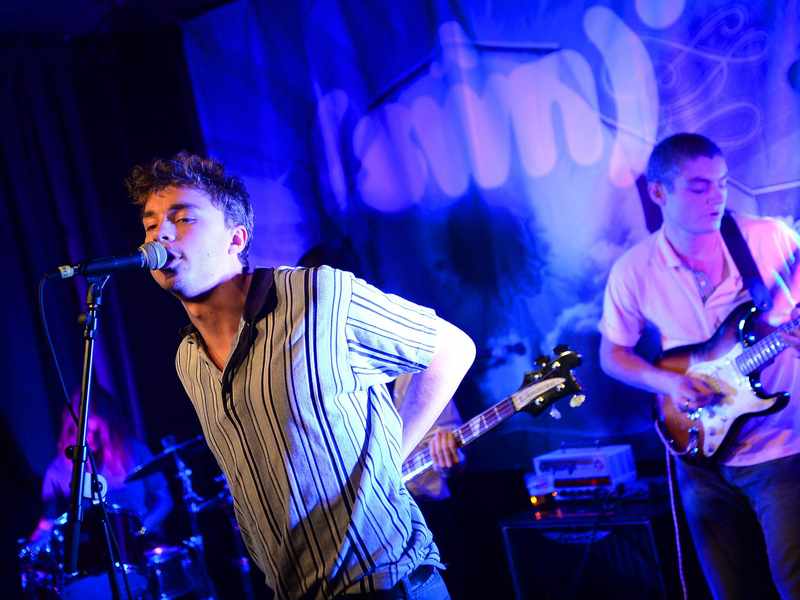 A person singing into a microphone. Behind them are a drummer and someone playing an electric guitar.