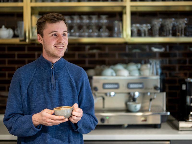 A person holding a coffee cup stands in a bar area. Behind them is a professional coffee machine.