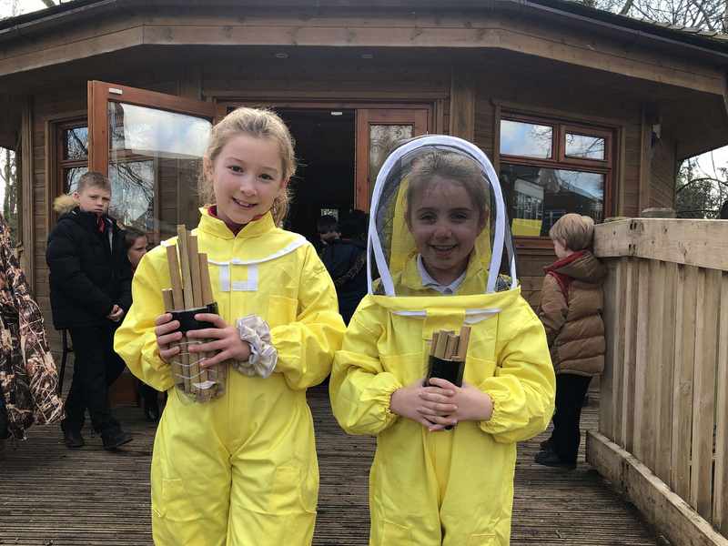 Two children wearing beekeeping suits stand in front of a small wooden building.