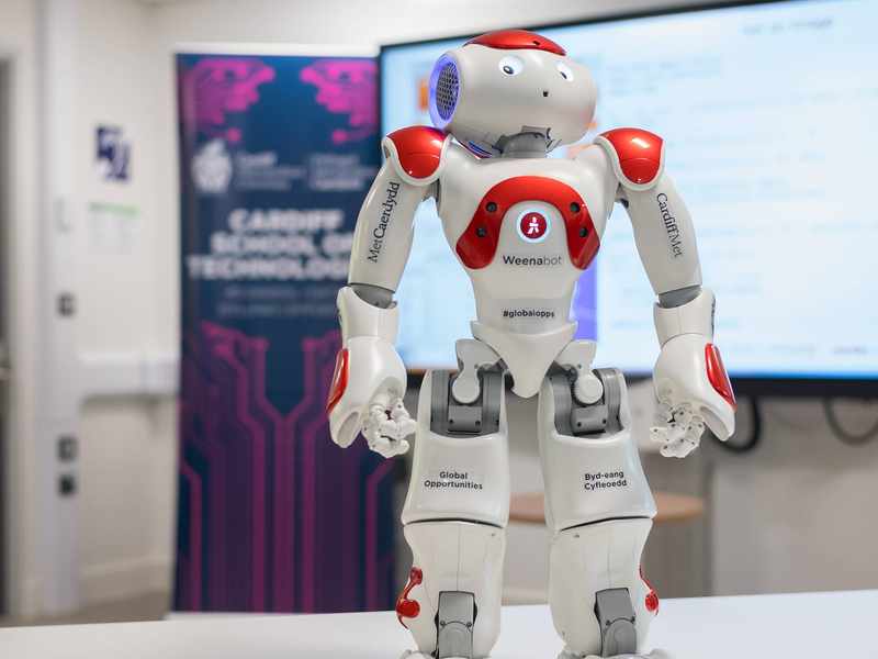 A close-up of a small humanoid robot. The robot has the Cardiff Met logo printed on its arms.
