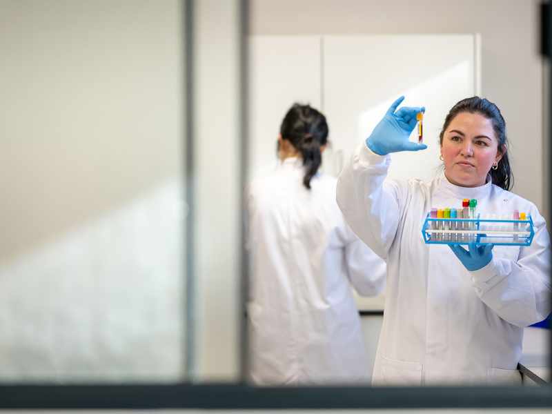 A female scientist in a white lab coat and blue gloves examines a test tube held up to the light. Another person in a lab coat works in the background. Various test tubes are organized in a rack. The scene is viewed through a glass window.