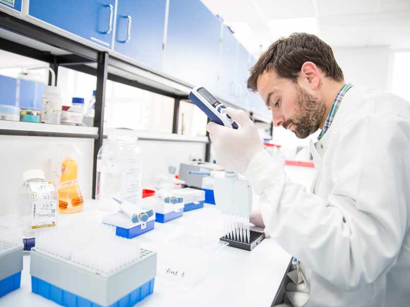 A scientist in a lab coat uses a pipette to transfer liquid into a tray, surrounded by lab equipment and bottles.