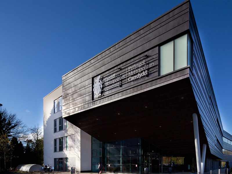 Modern building with angular architecture, featuring a large sign that reads Prifysgol Metropolitan Caerdydd. The structure has a mix of dark panels and white walls, with glass windows. Trees and a blue sky are visible in the background.