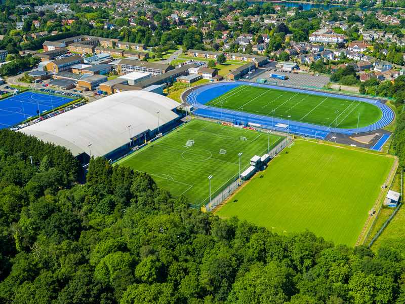 Aerial view of a sports complex featuring a large indoor sports facility, blue athletics track, multiple soccer fields, lush green trees in the foreground, and nearby residential buildings in the background.