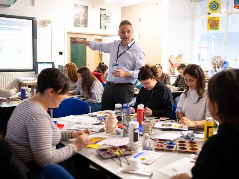 A teacher points at a whiteboard. He is surrounded by pupils at desks with paint and paper.